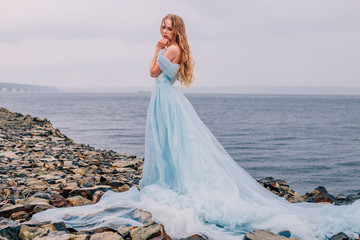 Beautiful young girl in a long blue lush dress is standing on the beach