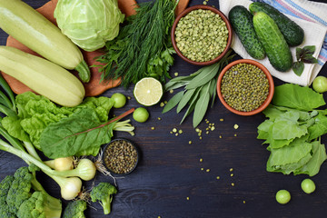 Composition on a dark background of green organic vegetarian products: green leafy vegetables, mung beans, zucchini, garlic, onions, cucumbers, peppers, lime. Top view. Flat lay.