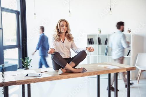 Fotografie, Obraz Middle aged businesswoman sitting on table and meditating in lotus position whil