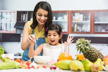 Pretty Indian young lady or mother with cute girl child or  daughter in kitchen having fun time with table full of fresh vegetables and fruits