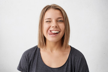 Wall Mural - Horizontal portrait of optimistic funny girl with bob hairstyle showing her tongue and blinking eyes while posing against white studio background. Emotional positive young female making grimace
