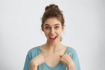 Wall Mural - Emotional woman with swept-up hair, blue eyes and white teeth being happy that she was chosen to be leader. Surprised beautiful joyful female with fair hair pointing at herself. Selective focus