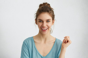 Wall Mural - Excited smiling young female with hair knot expressing positive emotions while posing against white background. Natural woman with pleasant wide smile wearing blue loose shirt raising her hand up