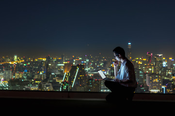 Wall Mural - Asian businessman sitting and using the laptop over the cityscape background at night time, Business success and technology concept