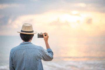 Wall Mural - Young Asian man traveler and photographer with jean shirt and hat taking photos of beautiful sunset at tropical beach island, background for summer holiday and vacation travel concepts