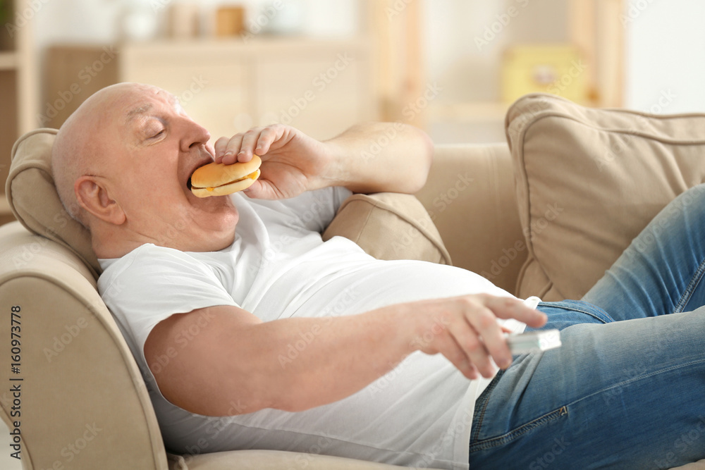 Fat senior man watching TV and eating hamburger while lying on sofa at home. Sedentary lifestyle concept