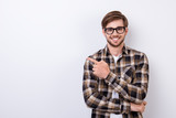 Fototapeta  - Smiling young nerdy bearded stylish student is standing on pure background in glasses and casual  outfit, pointing on the copyspace