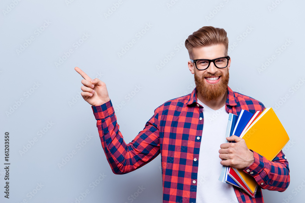 Smiling young nerdy red bearded stylish student is standing with books on pure background in glasses and casual bright outfit, pointing on the copyspace - obrazy, fototapety, plakaty 