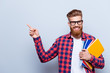 Smiling young nerdy red bearded stylish student is standing with books on pure background in glasses and casual bright outfit, pointing on the copyspace