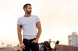 © Mikalai Bachkou - Sporty biker handsome rider male in white blank t-shirt walk away from classic style cafe racer motorbike at sunset. Vintage bike custom made in garage. Brutal urban lifestyle. Outdoor portrait.