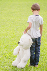 Wall Mural - Sad lonely boy with teddy bear standing in the meadow. Child looking down. Back view. Sadness, fear, loneliness concept