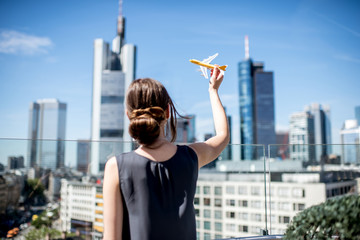 Wall Mural - Young woman playing with toy airplane on the modern cityscape background in Frankfurt. Air transportation concept in Frankfurt. Frankfurt has a very large airline connection in Europe