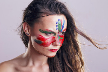 Girl with her face painted, colorful acrylic oil with her own hands, making random patterns. In studio, over a gray background.