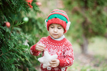Portrait Of A Little Boy In Elf Hat And Red Sweater Near The Christmas Tree And Holding Decoration