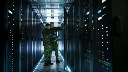 in data center two military men work with open server rack cabinet. one holds military edition lapto