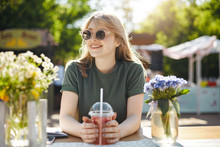 Young Beautiful Female Student Blogger Taking A Break From Classes Drinking Grapefruit Lemonade Smiling Looking Left Wearing Glasses In Park During A Food Festival On A Sunny Summer Day.