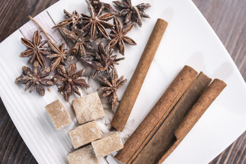 Three cinnamon sticks, anise stars and pieces of brown sugar lie on a white square saucer on a brown wooden background, top view