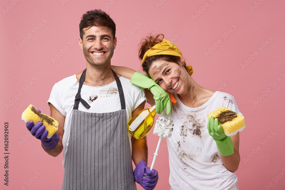 Housewife and her husband standing close to each other holding sponge, brush and cleaning solution having fun after cleaning room besmearing each other. Happy couple after cleaning their house - obrazy, fototapety, plakaty 