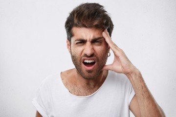 Portrait of depressed male with thick beard holding his hand on head having headache being sad after divorce with his wife. Emotional hipster guy looking troublesome frowning his face with displeasure