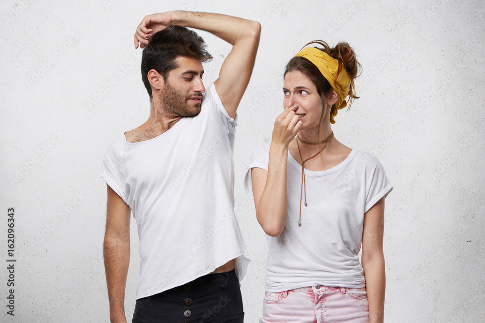 Female having disgusted look covering her nose with hand while smelling her husband`s armpit. Bearded man having nasty smell from sweat after running showing his wet underarm. Unpleasant smell - obrazy, fototapety, plakaty 