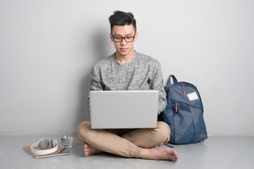 Young asian man in casual clothes is using a laptop, smiling while sitting on the floor