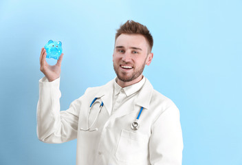 Poster - Happy young doctor holding piggy bank on light background