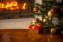 Closeup Image Of Red Gift Box On Wooden Table In Front Of Burning Fireplace And Christmas Tree. Empty Place For Text