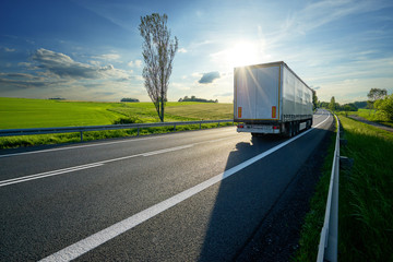 Truck driving on asphalt road along the green fields in rural landscape at sunset