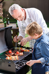 grey haired grandfather with his grandson preparing meat and vegetables on grill outdoors