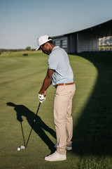 Wall Mural - Side view of african american man playing golf with club and ball at green lawn