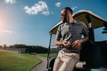 pensive man with golf ball in hand looking away while standing near golf cart