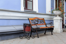 An Empty Wrought Iron Bench With Wooden Panels And Trash Can On The Background Of Blue Wall