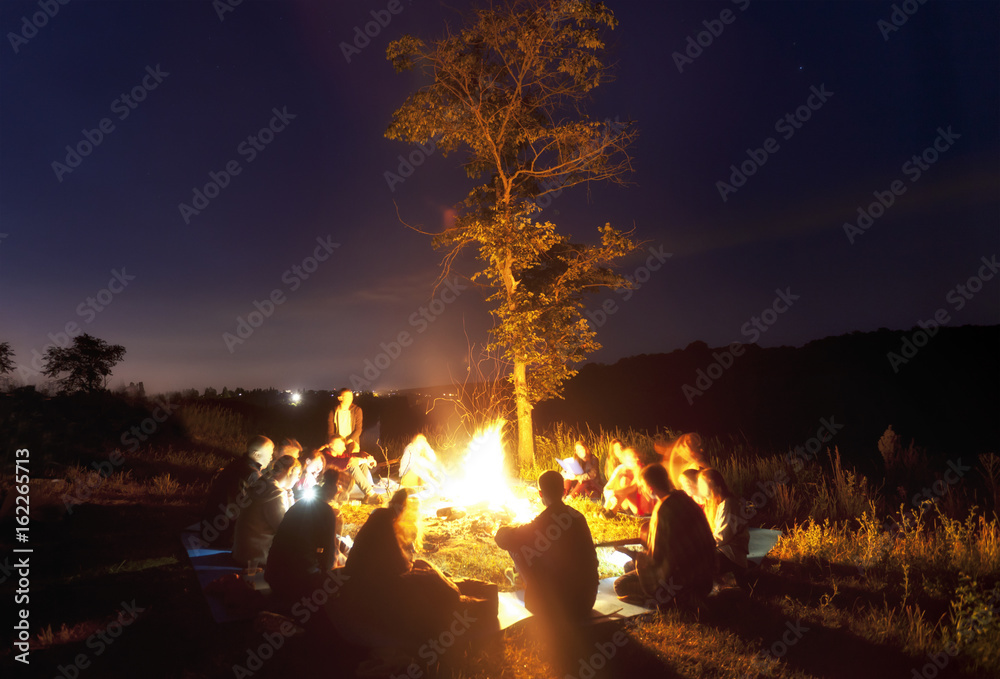 The company of young people are sitting around the bonfire and singing songs - obrazy, fototapety, plakaty 