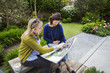 © Mint Images - Two women sitting on a bench in a garden, looking at a portfolio with gardening images, discussing garden design.