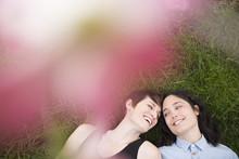 A Same Sex Couple, Two Women Lying On The Grass Laughing, Under The Branches Of A Flowering Tree. 