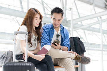 Couple asian student traveler looking smartphone and sit waiting for flight at airport terminal. Teenager are traveling concept.