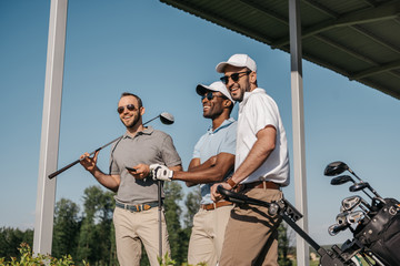 Wall Mural - Three smiling men in sunglasses holding golf clubs outdoors