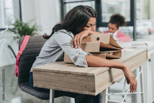 Fotografie, Obraz portrait of tired businesswoman lying on folders at workplace with daughter behi