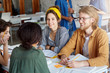 © WHstudio Leushin N - Back view of four students sitting together at table surrounded with books doing their common project sharing ideas with each other having pleasant smiles on their faces enjoying working together