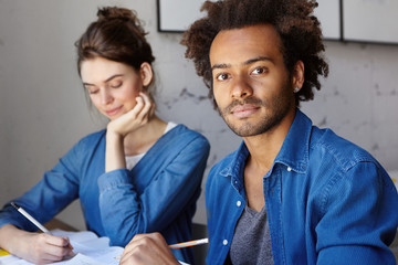 Wall Mural - Two students working on common research project sitting at desk in library, making notes in copybook. Confident dark-skinned A-student helping cute shy girl to solve difficult mathemtical problem