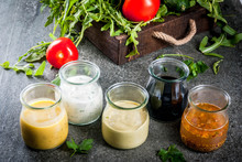 Set Of Dressings For Salad: Sauce Vinaigrette, Mustard, Mayonnaise Or Ranch, Balsamic Or Soy, Basil With Yogurt. Dark Stone Table. On Background Of Greenery, Vegetables For Salad. Copy Space