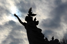Statue Of Girl Silhoetted By Pigeons With A Cloudy Sun Behind -making Her Look Like An Angel