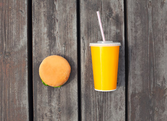 Fast food, unhealthy eating concept - juice cup and hamburger on a wooden table or floor background, top view