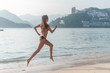 Back view of fit slim girl running barefoot on seashore wearing bikini. Young woman doing cardio exercise beach lit in sunshine and city mountains in background.