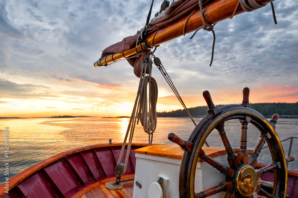 Sunrise sailing on a tall ship schooner.  Close up of steering wheel, bow and boom against a dramatic sky at dawn. - obrazy, fototapety, plakaty 