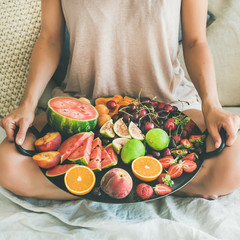 Summer healthy raw vegan clean eating breakfast in bed concept. Young girl wearing pastel colored home clothes sitting and holding tray full of fresh seasonal fruit, square crop