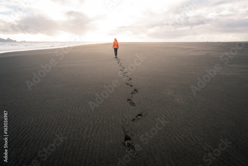 Fotomural Guy in Iceland walking on empty black sand beach into sunset leaving footprints