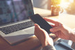 © oatawa - Hand of woman using mobile smartphone on wooden table with laptop in morning sunlight.
