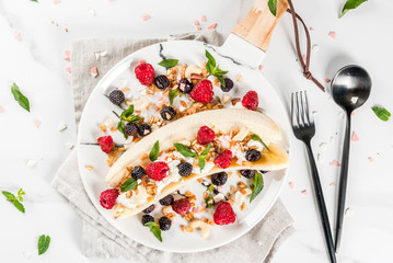 Summer fruit berry breakfast. Healthy banana split breakfast with cream cheese, raspberries, blackberries, mint, white and pink chocolate. On white marble plate, marble table. Top view copy space