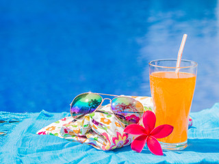 Wall Mural - Red frangipani (plumeria) flowers, sungrasses, shampoo, lotion, hat, rolled up towels and Orange juice with a piece of lemon at the side of swimming pool. Vacation, beach, summer travel concept